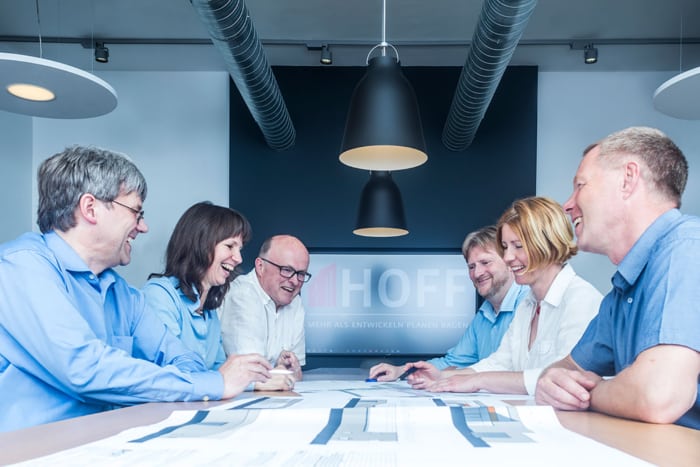 Sechs Personen sitzen lächelnd um einen Tisch in einem modernen Büro und diskutieren die vor ihnen ausgebreiteten Baupläne und Blaupausen. In dem hellen Raum sind Deckenleuchten und Lüftungsschächte zu sehen.
