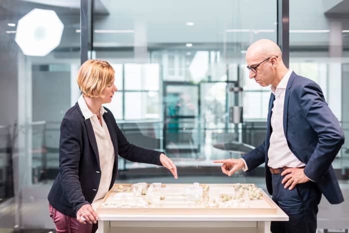 Zwei Fachleute, eine Frau und ein Mann in Geschäftskleidung, stehen an einem Tisch und besprechen ein Architekturmodell in einem modernen Büro mit großen Glasfenstern im Hintergrund.