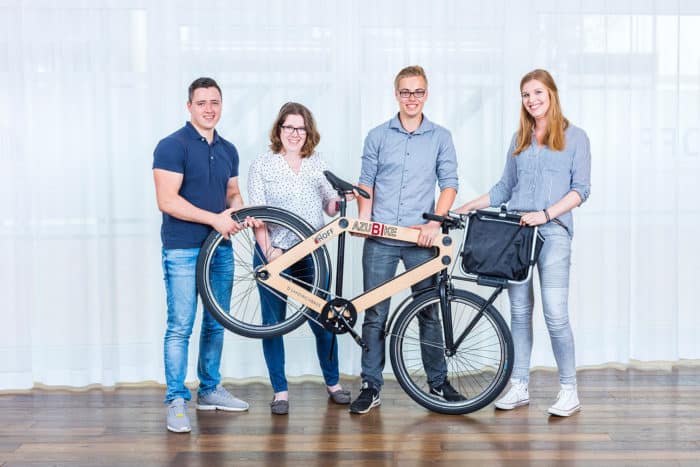 Vier lächelnde Menschen stehen drinnen und halten ein Fahrrad mit Holzrahmen. Zwei Personen halten das Fahrrad am Rahmen, eine andere hält den Lenker fest und eine weitere steht daneben. Sie sind leger gekleidet und stehen auf einem Holzboden.