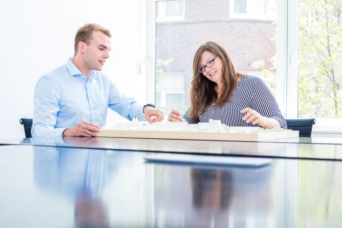 Zwei Personen sitzen an einem Tisch in einem hellen Büro und betrachten gemeinsam ein weißes Architekturmodell. Der Mann trägt ein hellblaues Hemd, die Frau eine Brille und eine gestreifte Bluse. Große Fenster lassen Tageslicht herein.