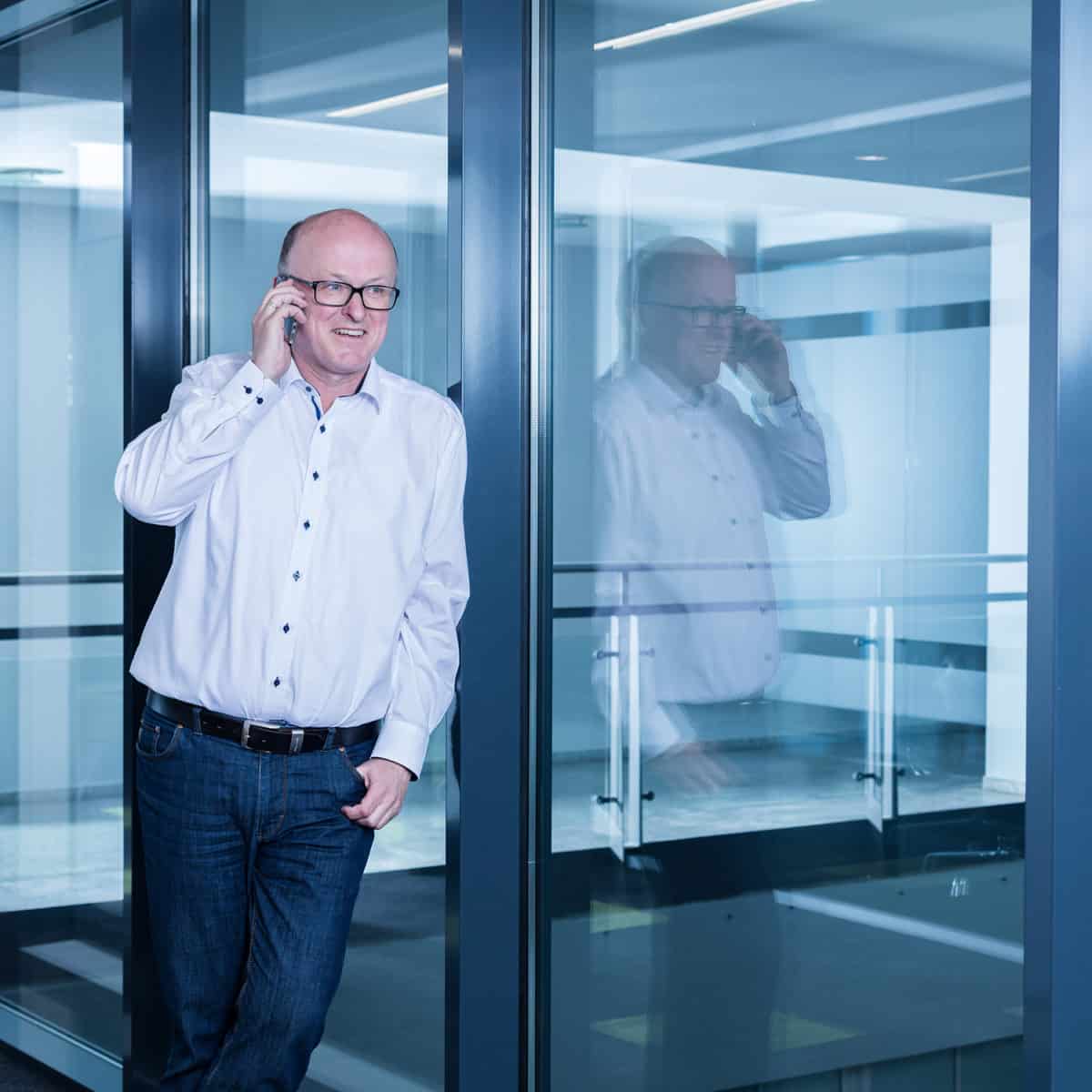 Ein Mann in weißem Hemd und Jeans steht an einer Glaswand und telefoniert. Sein Spiegelbild ist im Glas sichtbar, und er lächelt leicht in einer modernen Büroumgebung.