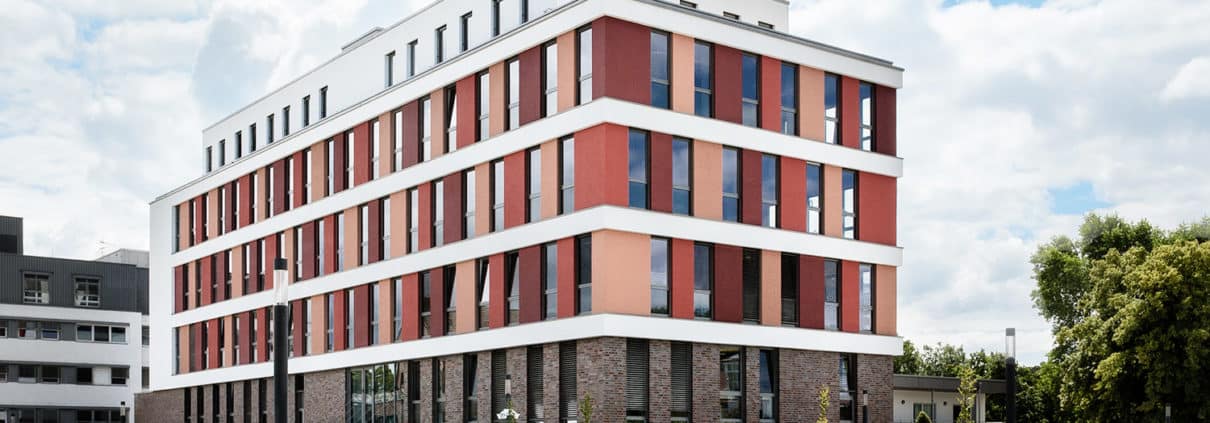 An einem leicht bewölkten Tag steht hinter einem vollen Parkplatz ein modernes, mehrstöckiges Gebäude mit rot-weißen und Backsteinfassaden. Bäume und gepflegte Büsche säumen den Parkplatz.