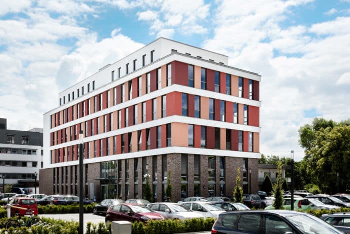 An einem leicht bewölkten Tag steht hinter einem vollen Parkplatz ein modernes, mehrstöckiges Gebäude mit rot-weißen und Backsteinfassaden. Bäume und gepflegte Büsche säumen den Parkplatz.