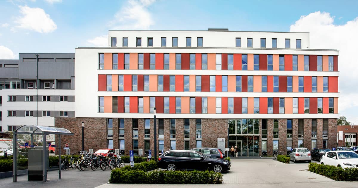 Ein modernes mehrstöckiges Krankenhausgebäude mit rot-weißen Paneelen, großen Fenstern und einem gemauerten Erdgeschoss, davor parken Autos und Fahrräder, unter einem blauen Himmel mit Wolken.