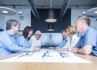 Sechs Personen sitzen lächelnd und unterhalten sich um einen Konferenztisch, vor ihnen liegen Dokumente in einem modernen Büro mit Hängelampen und freiliegenden Leitungen.
