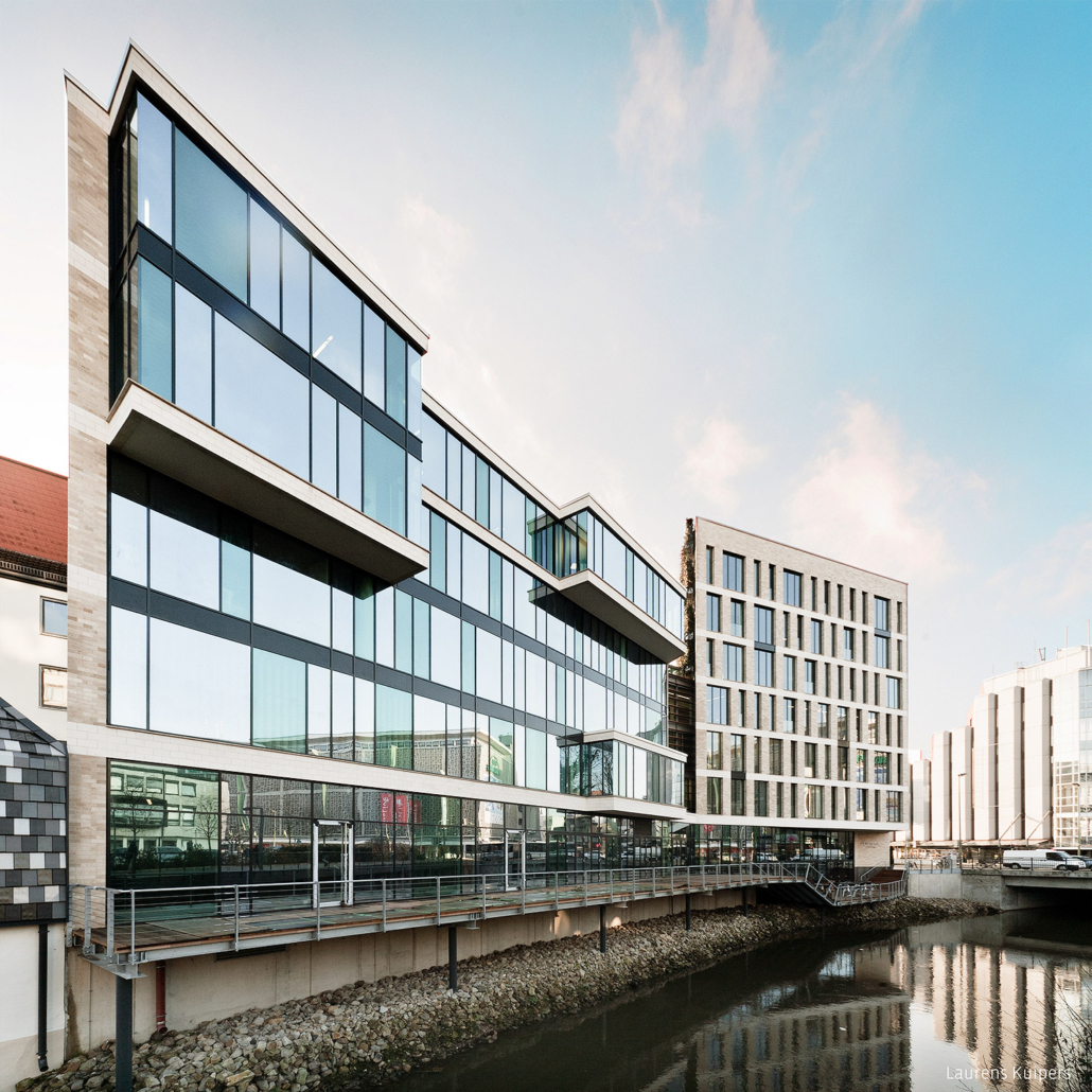 Ein modernes Bürogebäude mit großen Glasfenstern und geometrischer Architektur steht an einem Kanal, in dem sich der blaue Himmel und die Wolken spiegeln. Das Gebäude besticht durch Steinakzente und einen erhöhten Gehweg, der dem Wasser entlangführt.