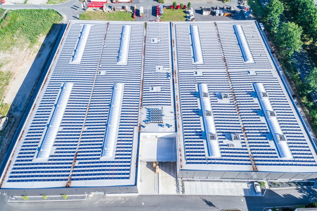 Luftaufnahme eines großen Industriegebäudes mit einem mit Solarmodulen bedeckten Flachdach, umgeben von gepflasterten Flächen, Grünflächen und Bäumen. Das Gebäude verfügt über mehrere Oberlichter und einen zentralen Eingang.