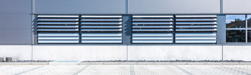 Eine moderne Gebäudefassade zeichnet sich durch große, horizontale Lamellenfenster aus Metall in einer grauen Wand über einem hellen Betonsockel aus, mit einer gepflasterten Fläche und kleinen dekorativen Steinen davor.