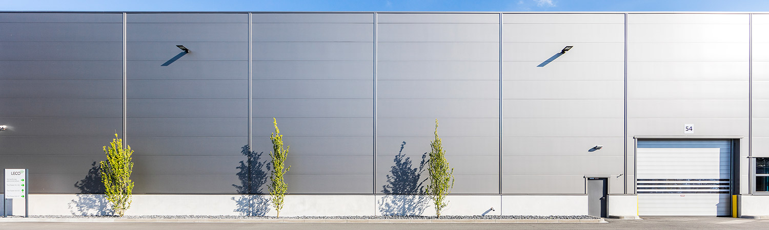 Ein modernes Industriegebäude mit einer flachen, grauen Metallfassade, drei jungen Bäumen und einem geschlossenen weißen Garagentor unter einem strahlend blauen Himmel.