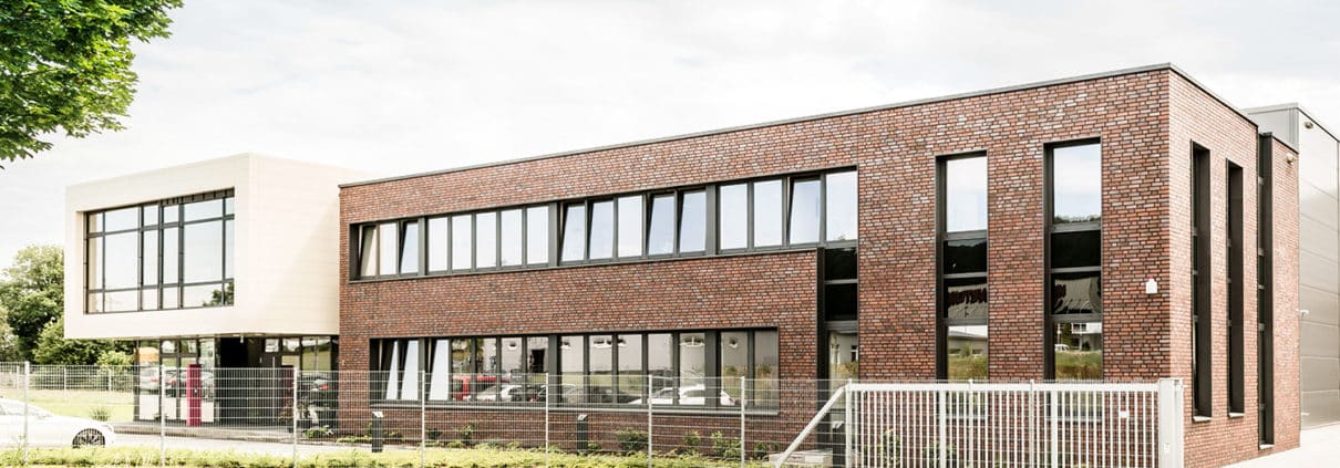Ein modernes, zweistöckiges Gebäude mit einer Mischung aus rotem Backstein und hellen Wandverkleidungen, großen Fenstern und einem Metallzaun davor, gelegen an einer geschwungenen Straße mit einem Baum und Grünflächen in der Nähe unter einem teilweise bewölkten Himmel.