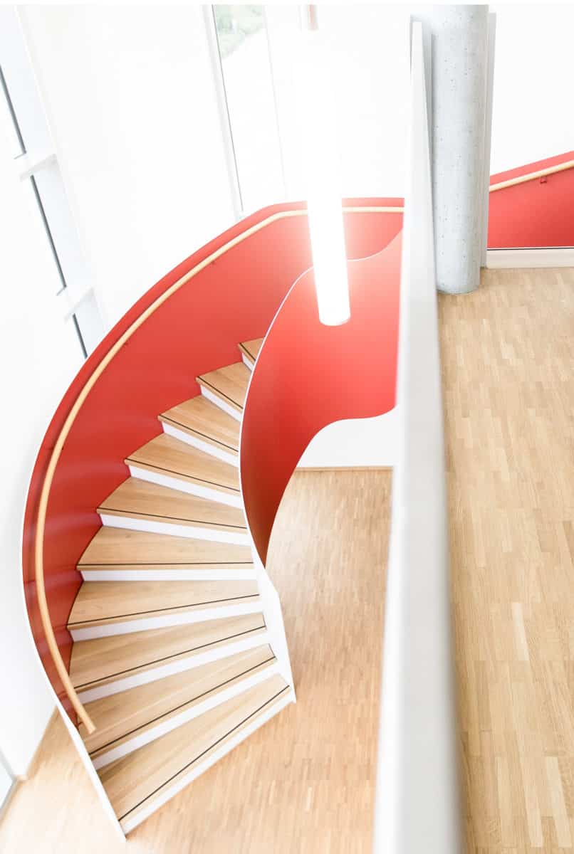 Eine Wendeltreppe mit roten Seiten und Holzstufen führt neben einer weißen Wand in einem hellen, modernen Innenraum mit großen Fenstern und hellem Holzboden nach oben.