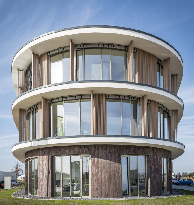 Ein modernes, rundes dreistöckiges Bürogebäude mit großen Fenstern, Holzvertäfelung und Ziegelsteinen im Erdgeschoss, vor einem blauen Himmel mit ein paar Wolken.