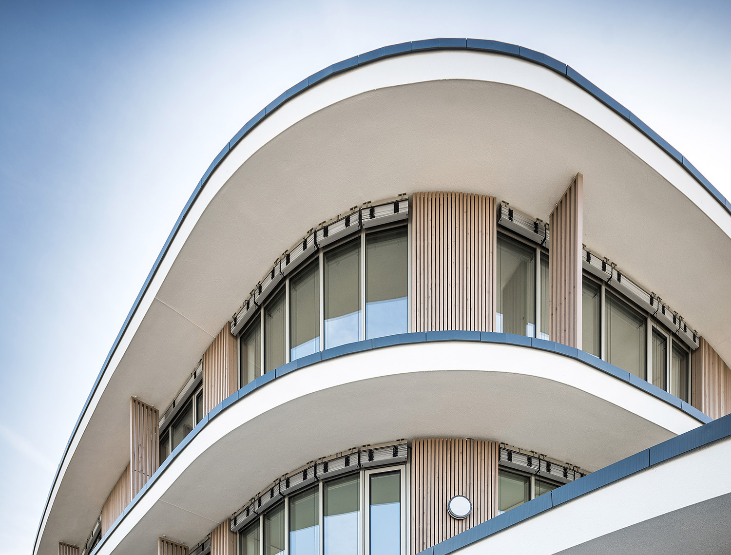 Modernes Bürogebäude mit glatten, geschwungenen Balkonen und großen Fenstern, mit vertikalen Holzlamellen und weißen Außenwänden, aus einem niedrigen Winkel vor einem klaren Himmel fotografiert.
