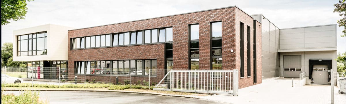 Ein modernes Industriegebäude mit einer Mischung aus Ziegeln und hellen Paneelen, großen Fenstern, einem Sicherheitszaun aus Metall und einer Einfahrt mit Tor unter einem teilweise bewölkten Himmel.