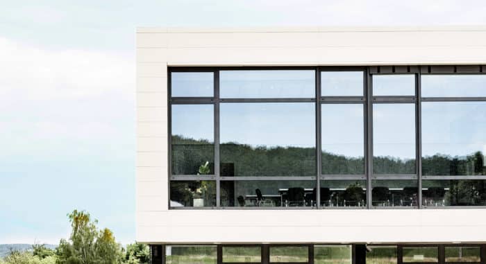 Ein modernes Gebäude mit großen, reflektierenden Fenstern, die Bäume und Himmel zeigen, im Inneren sind leere Bürotische und Stühle sichtbar. Die Außenfassade ist hell und von Grün umgeben.