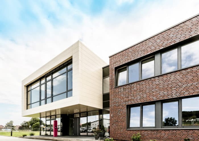 Ein modernes Gebäude mit großen Glasfenstern und einer Mischung aus beiger Verkleidung und roter Backsteinfassade unter einem teilweise bewölkten Himmel. In der Nähe des Eingangs sind Sträucher und ein rosa Schild zu sehen.