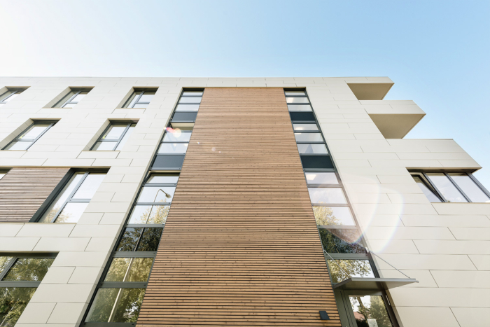 Modernes mehrstöckiges Gebäude mit weißen Außenwänden und vertikaler Holzvertäfelung, großen Fenstern, in denen sich Bäume spiegeln, und einem klaren blauen Himmel darüber.