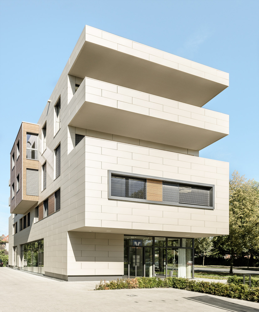 Ein modernes, mehrstöckiges Gebäude mit cremefarbenen rechteckigen Paneelen, großen horizontalen Überhängen und Glasfenstern im Erdgeschoss, umgeben von Grün und unter einem klaren blauen Himmel.