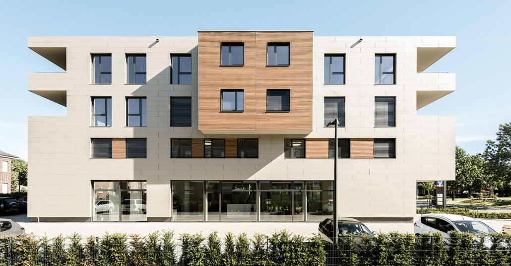 Ein modernes, vierstöckiges Gebäude mit einer Mischung aus beigem Stein und Holzpaneelfassade, großen Fenstern mit schwarzen Fensterläden, Glastüren im Erdgeschoss und Autos, die an einem sonnigen Tag hinter einer grünen Hecke davor geparkt sind.