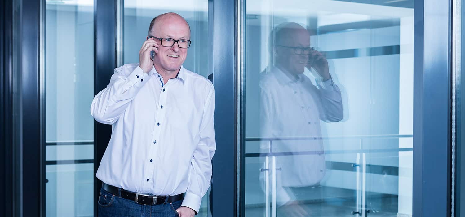 Ein Mann mit Brille und weißem Hemd steht in einem Innenbereich neben einer Glaswand und telefoniert mit seinem Smartphone. Sein Spiegelbild ist im Glas sichtbar.