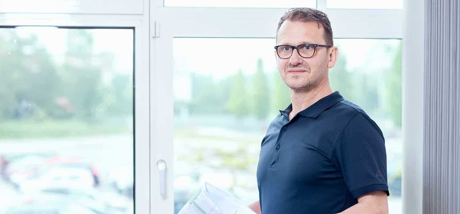 Ein Mann mit Brille und dunkelblauem Poloshirt steht mit Dokumenten in der Hand vor einem großen Fenster mit unscharfem Außenhintergrund.