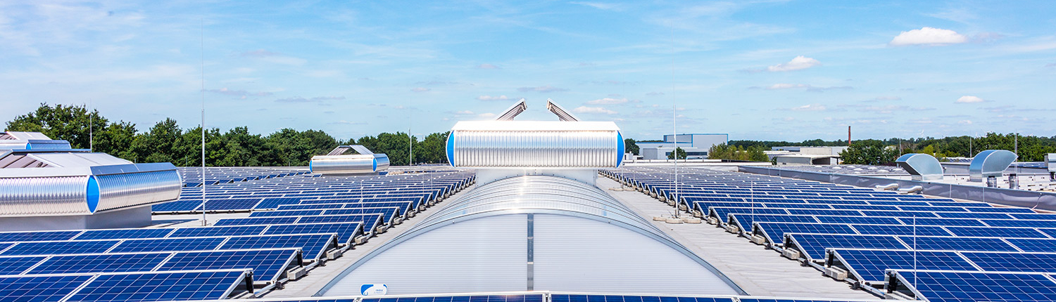 Auf einem großen Flachdach sind unter einem strahlend blauen Himmel Reihen blauer Solarmodule installiert, im Hintergrund sind Bäume und kleine Gebäude zu sehen.