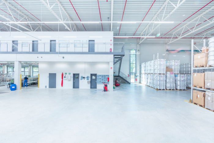 Ein geräumiges, modernes Lagerhaus mit hohen Decken, gestapelten Warenpaletten, großen Fenstern, einem oberen Zwischengeschoss und mehreren Türen entlang einer weißen Wand. Eine Treppe führt in das obere Stockwerk auf der rechten Seite.