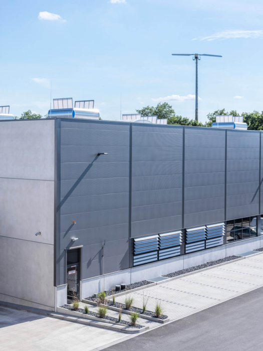 Ein modernes Industriegebäude mit grauen Wänden und wenigen Fenstern, gelegen in einem gepflasterten Bereich mit kleinen Pflanzen an der Seite. Der Himmel ist klar und blau, im Hintergrund sind Bäume zu sehen.