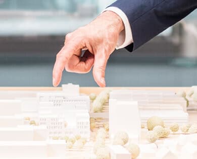 Eine Person im Anzug gestikuliert mit zwei Fingern über einem kleinen Architekturmodell mit weißen Gebäuden und runden Bäumen.