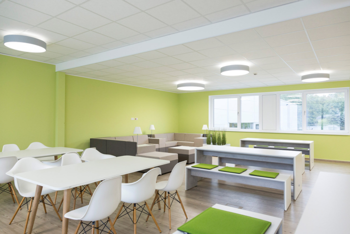 Ein moderner, heller Raum mit grünen Wänden, weißen Tischen, weißen Stühlen und Bänken mit grünen Kissen. Im hinteren Bereich stehen beige Sofas und große Fenster lassen viel Tageslicht herein.