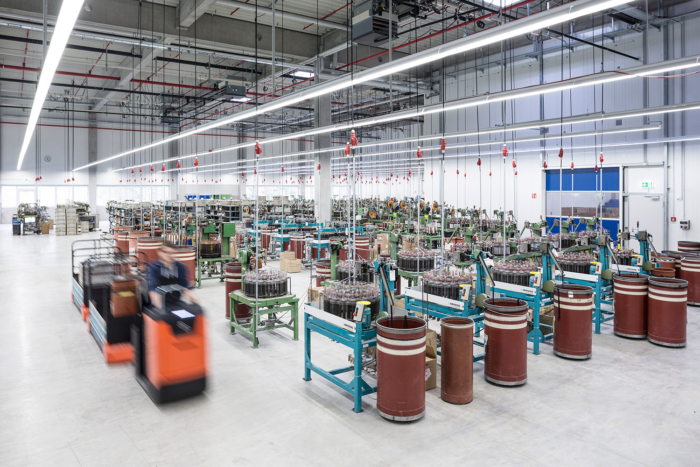 Eine geräumige, hell erleuchtete Fabrikhalle mit Maschinen und großen braunen Fässern in Reihen. Arbeiter bedienen Geräte und fahren orangefarbene Fahrzeuge. Über dem organisierten Arbeitsbereich sind Deckenrohre und Kabel sichtbar.