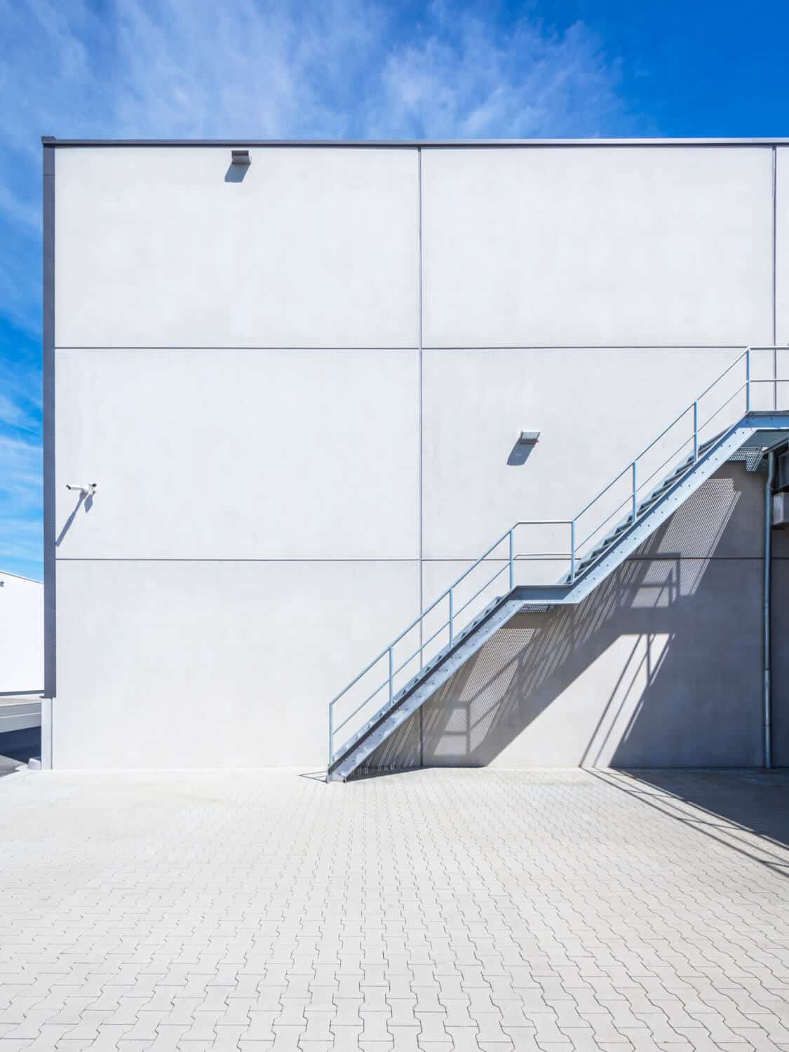Ein modernes Industriegebäude mit einer leeren weißen Wand, einer Metalltreppe im Freien, einer Überwachungskamera und einem strahlend blauen Himmel darüber. Der Boden ist mit hellen Ziegeln gepflastert.