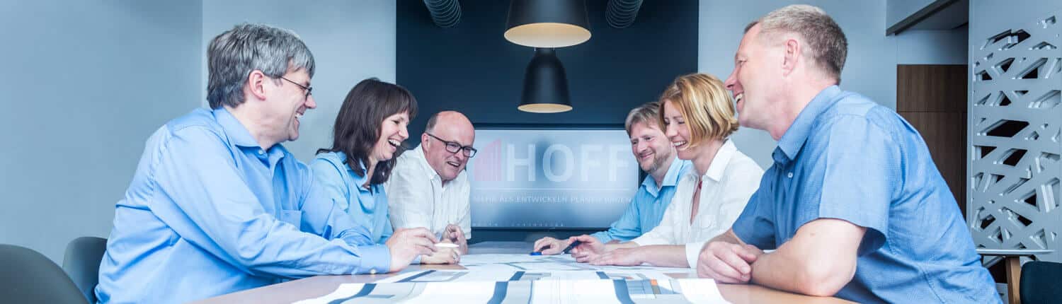 Sechs Teammitglieder in Geschäftskleidung sitzen lächelnd um einen Konferenztisch herum und arbeiten gemeinsam an Dokumenten in einem hellen, modernen Büro-Besprechungsraum.