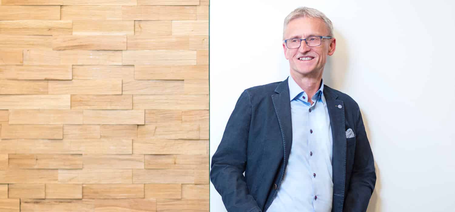 Ein Mann mit kurzen grauen Haaren und Brille, der einen blauen Blazer und ein helles Hemd trägt, steht lächelnd vor einer weißen Wand mit einem Holzpaneel-Akzent auf der linken Seite des Bildes.