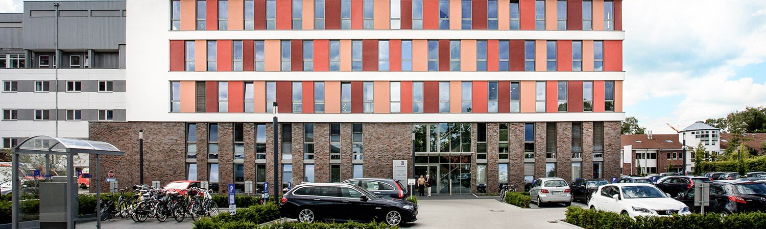 Ein modernes mehrstöckiges Gebäude mit einer Fassade aus Backstein und Glas, davor parkende Autos, Fahrräder auf der linken Seite und Grünflächen rund um den Eingang an einem teilweise bewölkten Tag.