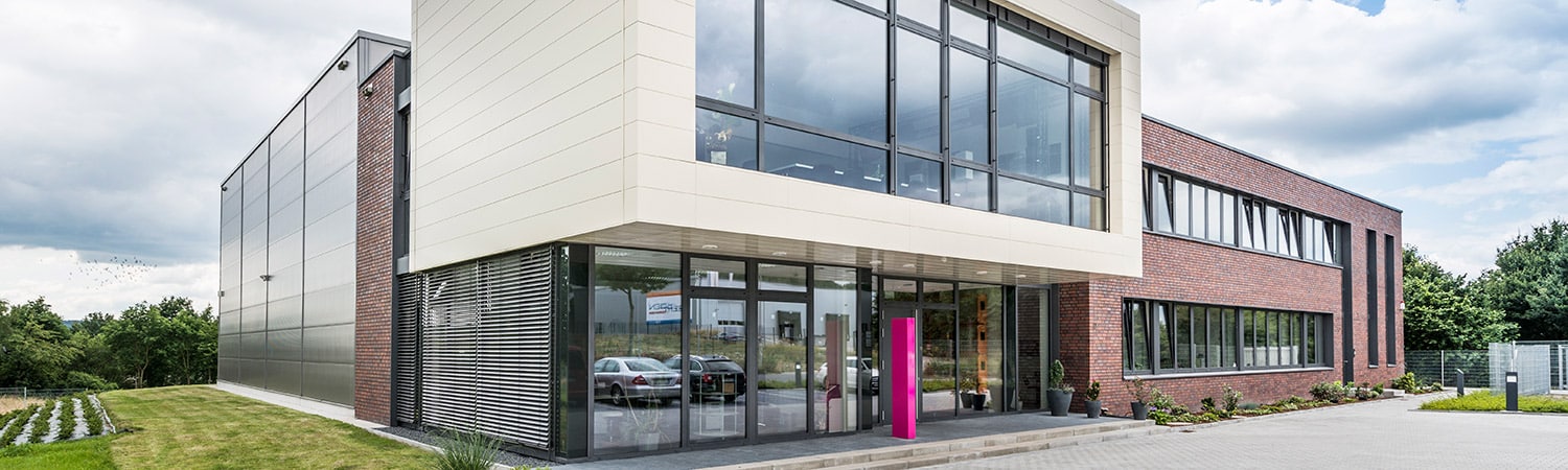 Modernes Geschäftsgebäude mit großen Glasfenstern, einer Fassade aus rotem Backstein und weißen Paneelen sowie einem begrünten Eingang. Durch die Fenster sind mehrere Autos sichtbar, und das Anwesen ist von Bäumen und Grün umgeben.