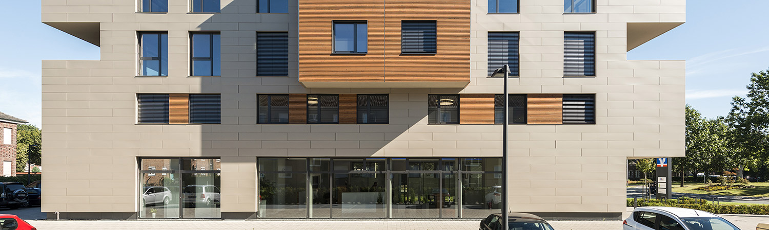 Moderne Gebäudefassade mit beigen Paneelen und Abschnitten aus horizontalen Holzlamellen, großen Glasfenstern im Erdgeschoss und davor parkenden Autos entlang der Straße. Im Hintergrund sind Bäume und blauer Himmel zu sehen.