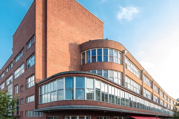 Ein großes Gebäude aus rotem Backstein mit geschwungenen und eckigen Abschnitten, breiten Fensterreihen und einem modernistischen Architekturstil vor einem blauen Himmel mit vereinzelten Wolken.