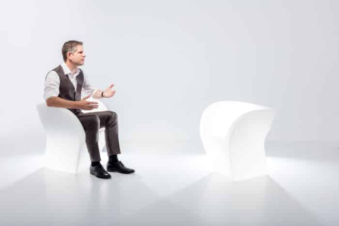Ein Mann in formeller Kleidung sitzt auf einem modernen weißen Stuhl und gestikuliert, als würde er mit jemandem sprechen. Ihm gegenüber steht in einem minimalistischen, hell erleuchteten weißen Raum ein identischer leerer Stuhl.