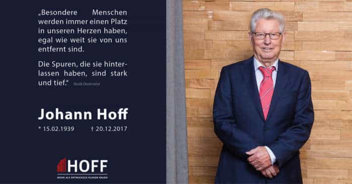 Ein älterer Herr in blauem Anzug und roter Krawatte steht lächelnd vor einer Holzwand. Neben ihm befindet sich ein Gedenktext für Johann Hoff (1939–2017), der sein Andenken und sein Wirken würdigt.