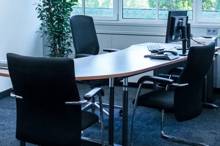 Ein modernes Büro mit einem geschwungenen Schreibtisch, drei schwarzen Stühlen, einem Computermonitor, einer Tastatur und einer Maus. In der Ecke steht eine hohe grüne Pflanze, und große Fenster lassen Tageslicht herein.