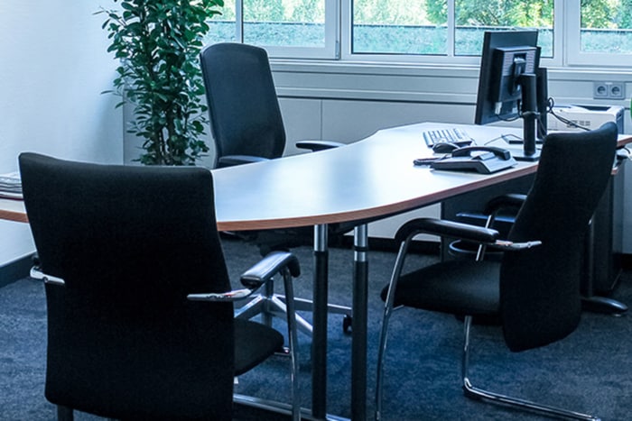 Ein modernes Büro mit einem geschwungenen Schreibtisch, drei schwarzen Stühlen, einem Computermonitor, einer Tastatur und einer Maus. In der Ecke steht eine hohe grüne Pflanze, und große Fenster lassen Tageslicht herein.