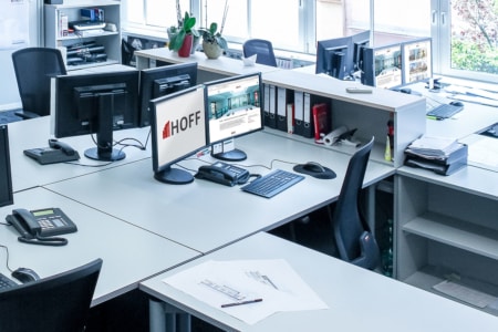 Ein modernes Büro mit mehreren Schreibtischen, Computern, Telefonen, Bürostühlen, Ordnern und Papieren. Auf einem Computerbildschirm steht das Wort „HOFF“. Große Fenster sorgen für Tageslicht. Der Arbeitsbereich wirkt aufgeräumt und sauber.