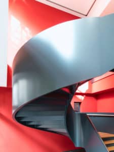 Eine moderne Wendeltreppe mit glatten, grauen Metallstufen und Geländer bildet einen Kontrast zu den leuchtend roten Wänden, die durch natürliches Licht von oben beleuchtet werden.