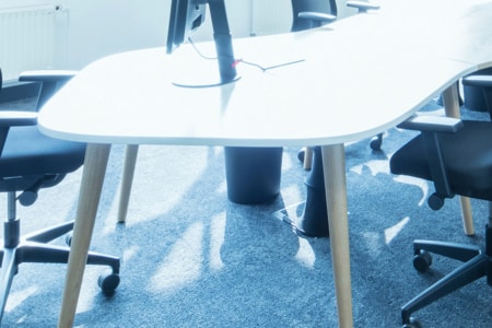 Ein moderner Bürotisch mit hellen Holzbeinen, umgeben von schwarzen Bürostühlen auf blauem Teppich. Ein Computermonitor steht auf dem Tisch. Helles Tageslicht durchflutet den Raum.