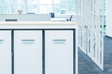 Moderner Büroraum mit weißen Schränken im Vordergrund, einer dekorativen weißen Trennwand auf der rechten Seite, grauem Teppichboden und großen Fenstern im Hintergrund, die natürliches Licht hereinlassen.