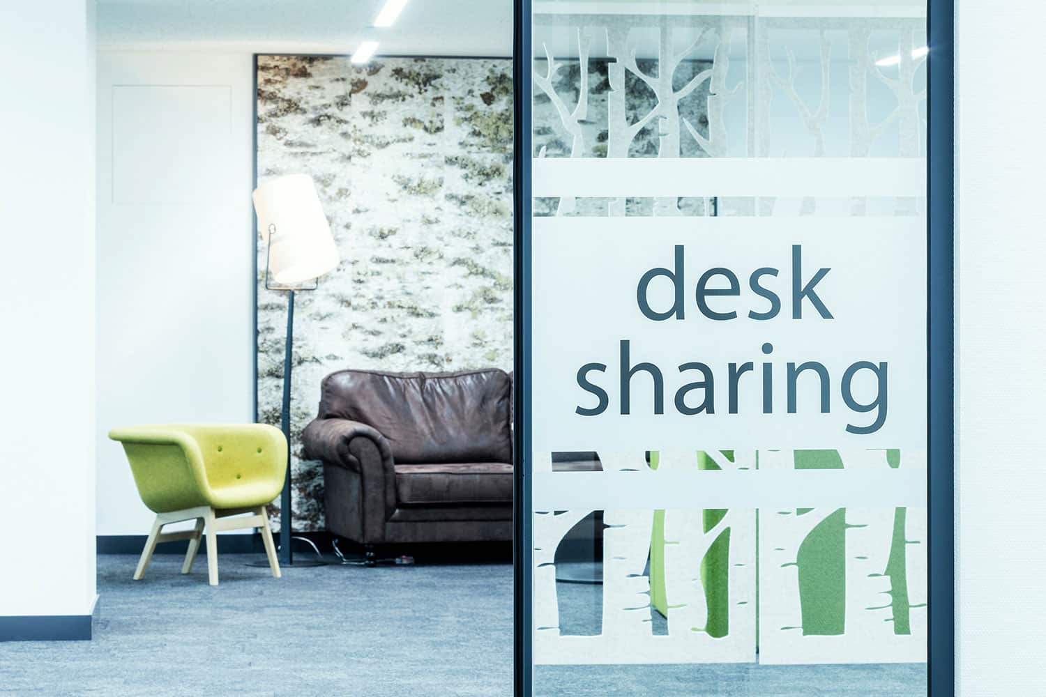 Ein moderner Büroraum mit einer Glastrennwand mit der Aufschrift „Desk Sharing“, einem braunen Ledersofa, einem grünen Sessel, einer Stehlampe und einer Wand mit Naturmotiven.