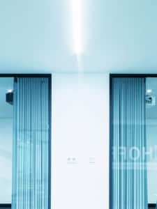 Minimalistisches, modernes Interieur mit weißen Wänden, zwei hohen Glastüren mit blauen Vertikaljalousien und einer hellen Deckenleuchte. Der Raum wirkt sauber und leer, mit schwachen Reflexionen auf dem Glas.