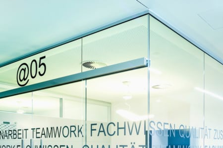 Ein modernes Bürointerieur mit Glaswänden und dem Schriftzug @05 am oberen Ende. Auf den Glasplatten sind Worte wie TEAMWORK und FACHWISSEN aufgedruckt. Deckenleuchten und Lüftungsöffnungen sind im Inneren sichtbar.
