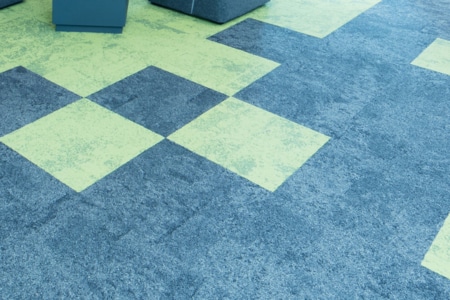 Ein Teppichboden mit abwechselnd grünen und blauen quadratischen Fliesen in einem geometrischen Muster, wobei im oberen Bereich ein Teil einer grau gepolsterten Sitzfläche sichtbar ist.